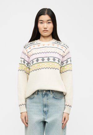 Tânără cu păr lung și închis la culoare, purtând un pulover crem cu modele pastelate de inimioare și semne ale păcii, asortat cu blugi din denim albastru deschis.