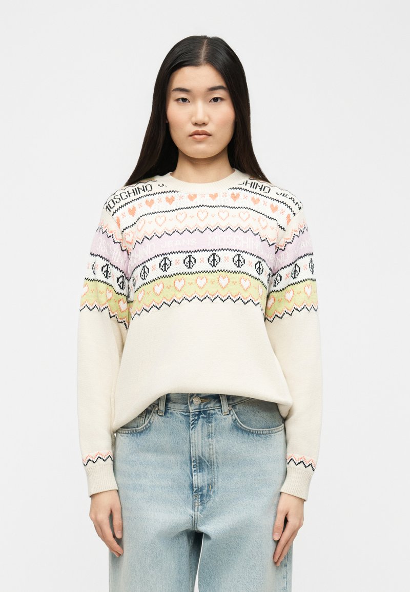 Νεαρή γυναίκα με μακριά σκούρα μαλλιά, φορώντας ένα εκρού πουλόβερ με παστέλ σχέδια καρδιάς και σήμα ειρήνης, συνδυασμένο με ανοιχτόχρωμα τζιν.