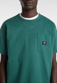 Camiseta de algodón verde con cuello redondo, mangas cortas y un bolsillo en el pecho izquierdo con una etiqueta negra de Vans. Textura suave.