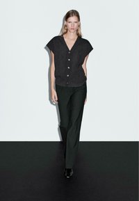 Top in maglia corto a maniche con scollo a V e dettaglio di bottoni grigio scuro, abbinato a pantaloni slim-fit verdi scuri e stivaletti neri alla caviglia.