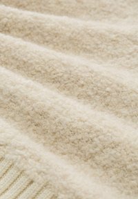 Beżowa tkanina o fakturze z miękką, puszystą powierzchnią i prążkowanym brzegiem. Materiał wydaje się ciepły i przyjemny, z delikatnie falującymi falami.