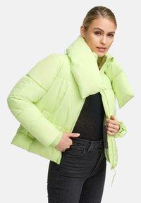 Vrouw in een lichtgroene pufferjack met een grote kraag, zwarte top en donkere high-waisted jeans tegen een effen achtergrond.
