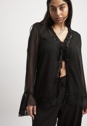 Black sheer blouse featuring lace trim, ruffled edges, and tie closure. Long sleeves with flared cuffs; smooth texture.