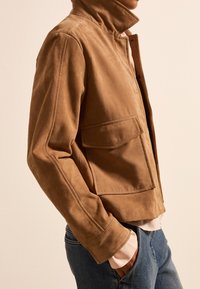 Braune Wildlederjacke mit hohem Kragen, seitlichen Taschen und tailliertem Design, kombiniert mit blauen Jeans. Glatte Textur und minimalistische Details.