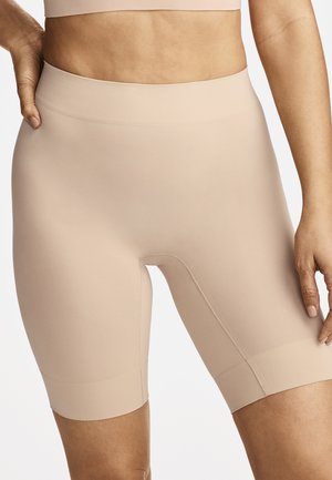 Woman wearing high-waist, beige, seamless biker shorts and matching sports bra, standing with one hand on hip against a plain background.