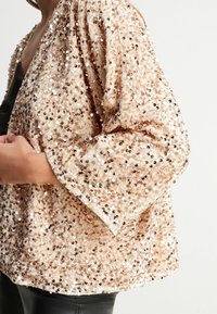 Veste couverte de sequins en beige clair, dotée d'une coupe ample et de manches larges. Le tissu présente une texture avec une surface scintillante et réfléchissante.