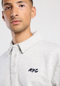 Polo shirt grigio chiaro con colletto pieghevole e tre bottoni, con ricamo blu navy "NYC" sul lato sinistro del petto. Tessuto morbido.