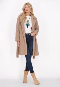 Cappotto beige, morbido e lungo con rever, indossato sopra una maglietta bianca e jeans scuri. Collana piumata notevole e stivali alla caviglia color sabbia chiara.