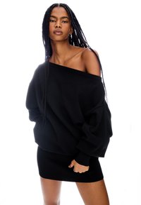 Överstor svart tröja med off-shoulder-design, kombinerad med en ribbad svart minikjol. Texturerad tyg, lös passform och långa ärmar.
