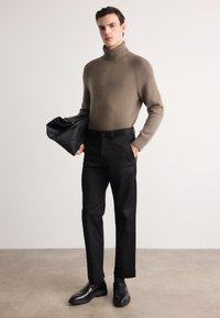 Brun strikket turtleneck-sweater kombineret med sorte skræddersyede bukser. Modellen holder en sort lædertaske og bærer sorte sko.