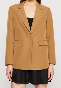 Hnědý blazer s přiléhavým střihem, výřezovými klopami, zapínáním na jeden knoflík a dvěma předními kapsami, doplněný černými šaty pod ním.