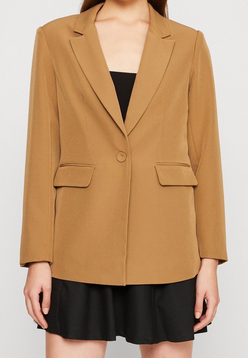 Hnědý blazer s přiléhavým střihem, výřezovými klopami, zapínáním na jeden knoflík a dvěma předními kapsami, doplněný černými šaty pod ním.