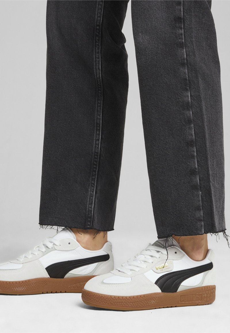 Personne portant des baskets blanches Puma avec des bandes noires et des semelles en gomme, associées à un jean foncé à ourlet effiloché, debout sur une surface grise.