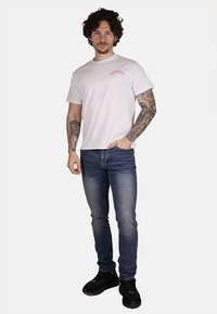 T-shirt bianca in cotone con piccolo logo rosso, abbinata a jeans blu scuri. Il modello indossa sneakers nere e si trova in piedi con le mani nelle tasche.