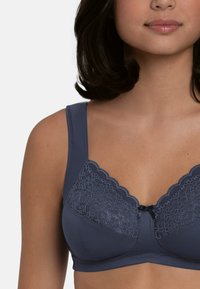 Grijze bh met een getextureerd kant paneel, geschulpte randen, brede bandjes en een klein decoratief strikje in het midden. Soepele stofafwerking.