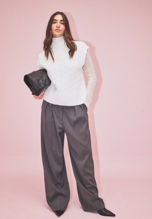 Pull en maille blanche épaisse sans manches, associé à un pantalon large taille haute gris foncé. Talons aiguilles noirs et pochette.