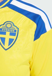 Gelbes Sporttrikot mit blauen Akzenten, versehen mit einem strukturierten Wappenemblem und dem Schriftzug "SVERIGE" in Blau. Weiß gestreiftes Schulterdesign.