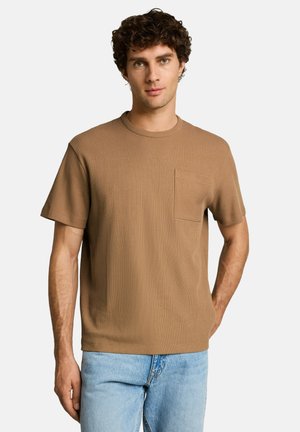 Man met krullend haar die een bruin geribbeld shirt met korte mouwen en borstzakje draagt, en lichtblauwe spijkerbroek, staand tegen een witte achtergrond.