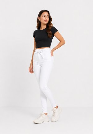 Frau trägt ein schwarzes Crop-T-Shirt, hoch taillierte weiße Hose mit einer Schleifengürtel und weiße klobige Sneaker, die vor einem einfarbigen hellen Hintergrund steht.