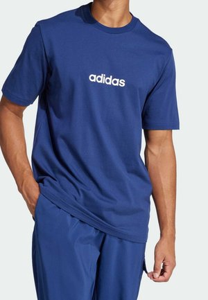 Mężczyzna ubrany w niebieską koszulkę Adidas z białym logo na środku klatki piersiowej, w połączeniu z pasującymi niebieskimi spodniami, ręka spoczywająca w kieszeni.