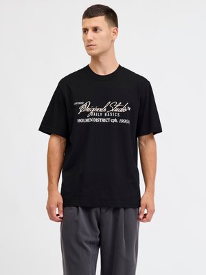 Junger Mann trägt ein schwarzes, übergroßes T-Shirt mit weißem Text und graue Hosen, steht vor einem schlichten weißen Hintergrund.