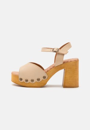 Beżowa sandałka z zaokrąglonym noskiem, otwartym bokiem, regulowanym paskiem na kostkę oraz obcasem z korkopodobnego drewna. Ozdobiona dekoracyjnymi ćwiekami.