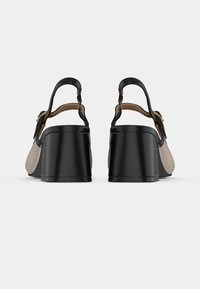 Sandales à talons hauts noires et beiges avec un talon épais, une bride à la cheville et des détails en métal doré. Texture en cuir lisse avec un design contrasté.