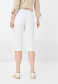 Pantaloni capri bianchi con un design aderente, dotati di tasche posteriori e orli arrotolati, abbinati a espadrillas beige chiare su uno sfondo minimalista.