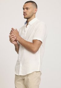 Chemise en lin blanc à manches courtes avec un col boutonné, présentant une texture subtile et un accent bleu clair à l'intérieur du col.