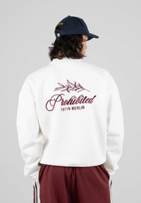 Persona con capelli ricci scuri, che indossa un cappellino blu navy, una felpa bianca con il testo "Prohibited 10119 Berlin" e pantaloni bordeaux con strisce bianche.