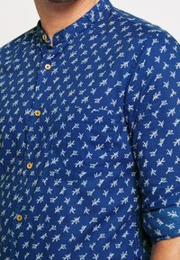 Blå skjorta med mandarinkrage, vita bladliknande mönster, träknappar och en bröstficka, med en texturerad tygfinish.