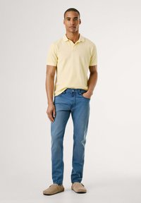 Polo majica v svetlo rumeni barvi, modri kavbojki in bež natikačih. Prilagojena zasnova z mehko teksturo. Model stoji z rokami v žepih.