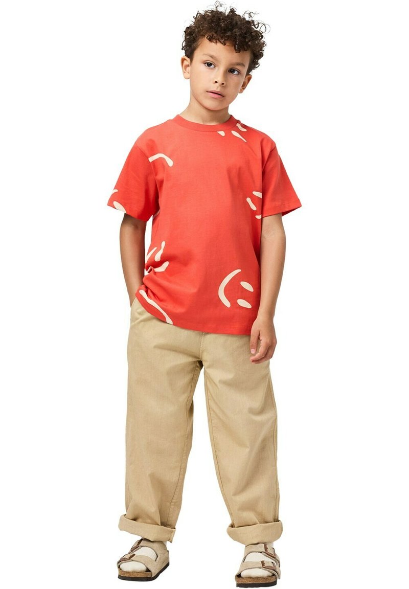 T-shirt rouge en coton avec des formes abstraites blanches, associé à un pantalon ample beige et des sandales marron. Design simple, coupe décontractée.