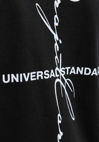 Černá látka se bílým potiskem, na kterém je nápis "UNIVERSAL STANDARD" a zakřivené akcenty. Textura vypadá hladce a jemně.