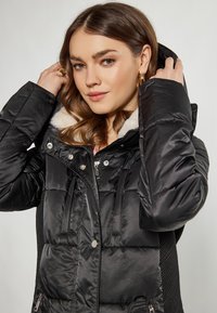 Jachetă puffer neagră, cu o glugă moale căptușită cu blană albă, buzunare cu fermoar și o textură matlasată cu accente lucioase.