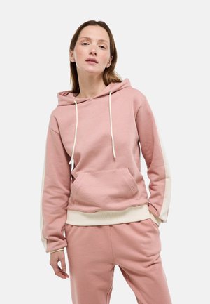 Femme portant un sweat à capuche rose poussiéreux et un pantalon de jogging assorti avec des rayures crème et des poignets, debout devant un fond clair uni.
