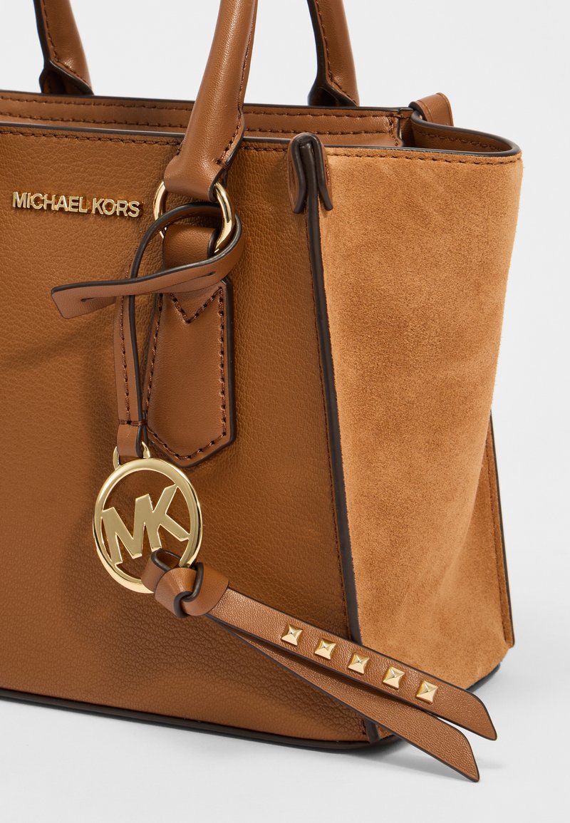 Sac en cuir marron avec des accents en daim, ornée de quincaillerie dorée, d'un charm logo circulaire et de clous en pyramide sur une bandoulière.