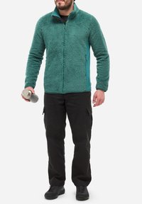 Mann trägt eine türkisfarbene Fleecejacke, schwarze Cargo-Hosen und schwarze Schuhe, hält in seiner linken Hand eine silberne isolierte Wasserflasche.