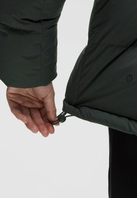 Tumeda roheline puhvjakot, millel on nähtav köis ja lukk. Käsi hoiab köiest, näidates kangast tekstuuri ja tikandeid.