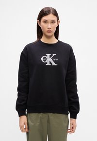 Čierna mikina s okrúhlym výstrihom, pružnými manžetami a veľkým strieborným logom 'CK'. Vyrobená z hladkej, mäkkej látky, s voľným strihom.