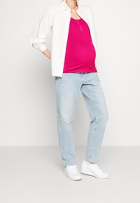 Těhotenský outfit s růžovým žebrovaným tílkem, bílou košilí na knoflíky, světle modrými džíny a bílými teniskami. Přiléhavý, ležérní styl.