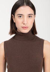 Top senza maniche in maglia liscia di colore marrone, con colletto alto e texture a costine sul collo, dal design aderente.