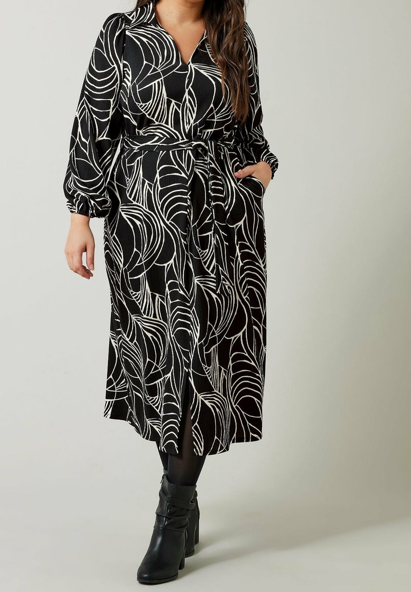 Femme portant une robe midi noire avec des motifs abstraits blancs en forme de feuilles, des manches longues bouffantes, une ceinture à la taille, des collants noirs et des bottines noires à talons.