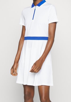 Vrouw draagt een witte jurk met korte mouwen, een blauwe kraag en een blauwe tailleband, staand tegen een effen lichte achtergrond.