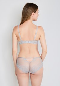 Femme avec de longs cheveux bruns portant un ensemble de lingerie en dentelle gris clair, se tenant de dos face à un fond uni clair.