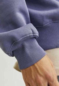 Nahaufnahme einer Hand, die einen lila gerippten Pulloverärmel über einem beigen Hemd trägt.