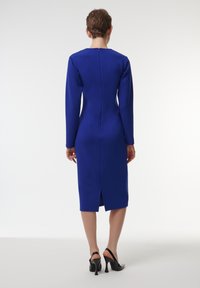 Vestido azul de manga larga con cierre en la parte posterior, diseño ajustado, hasta la rodilla y con una abertura en la parte trasera. Modelo lleva puestos zapatos de tacón negro.