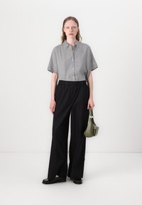 Gestreept short-sleeve shirt met knopen, gecombineerd met hoge zwarte wijde broek. Model houdt een kleine groene tas vast.