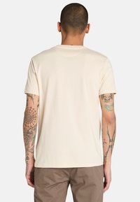 Beige tričko s krátkým rukávem vyrobené z měkké bavlny. Design s kulatým výstřihem a hladkou texturou. Viditelné tetování na pažích.