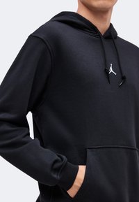 Personne portant un sweat à capuche noir avec une poche frontale et le logo Jumpman blanc, main dans la poche, visible de la poitrine à la taille.